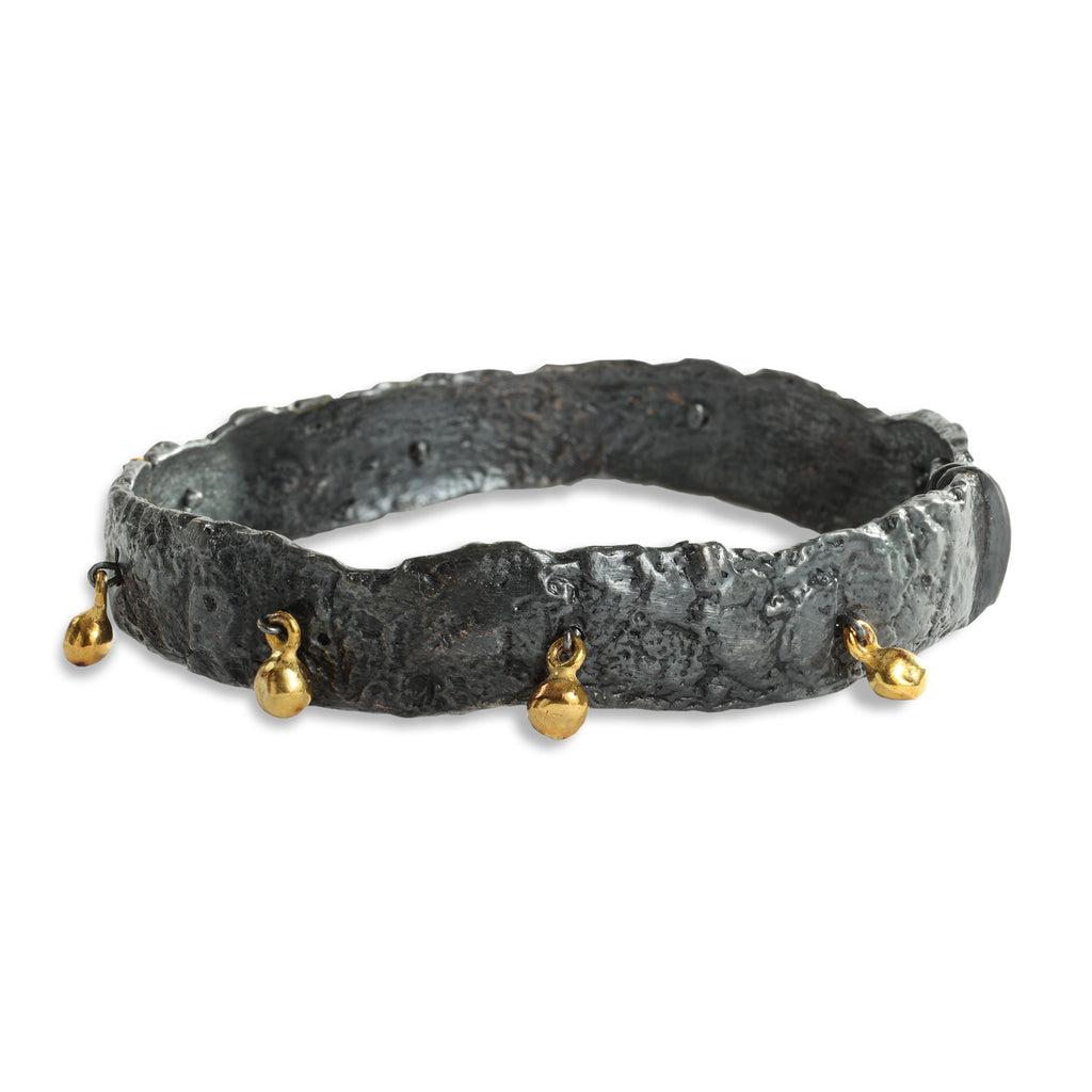 This Svelare bangle in blackened sterling silver, showcases a captivating array of dangling textured elements crafted in 18k yellow gold. As it adorns the wrist, the wearer is treated to the delightful sound of the gold elements gently striking against the blackened silver. This bold contrast of materials creates a striking aesthetic.  • Blackened sterling silver, 18k yellow gold  • Clasp