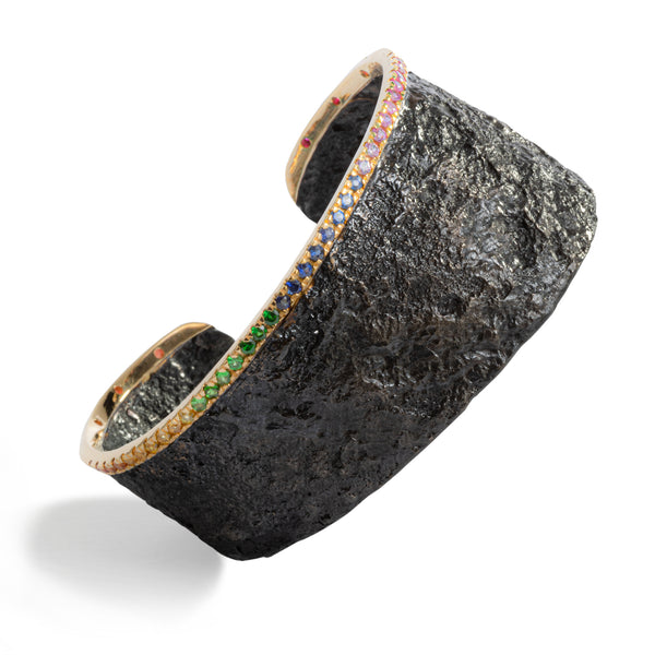The November's Moon cuff showcases an intriguing design, featuring an 18k yellow gold bar that sets multicolor sapphires in a rainbow-inspired arrangement over highly textured blackened sterling silver. With its asymmetrical cuff, this piece embodies a sleek, earthy combination that speaks volumes of personality.  • Sapphire ct: 0.86