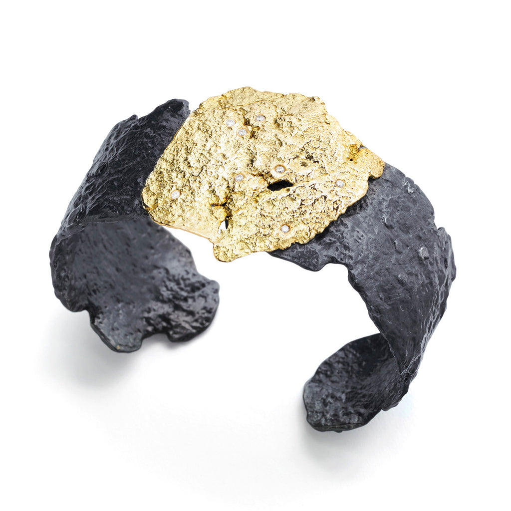 Blackened Silver 18k Gold Diamond Roccia Cuff Bracelet by Emanuela Duca. The ragged surfaces of 18k yellow gold and blackened sterling silver create a primal fusion of past and present, where the timeless seamlessly meets the contemporary. Within this lies 8 white diamonds, 1.5mm each. The imperfections and empty space on the surfaces evoke a sense of unearthed treasures, hinting at a story buried within.